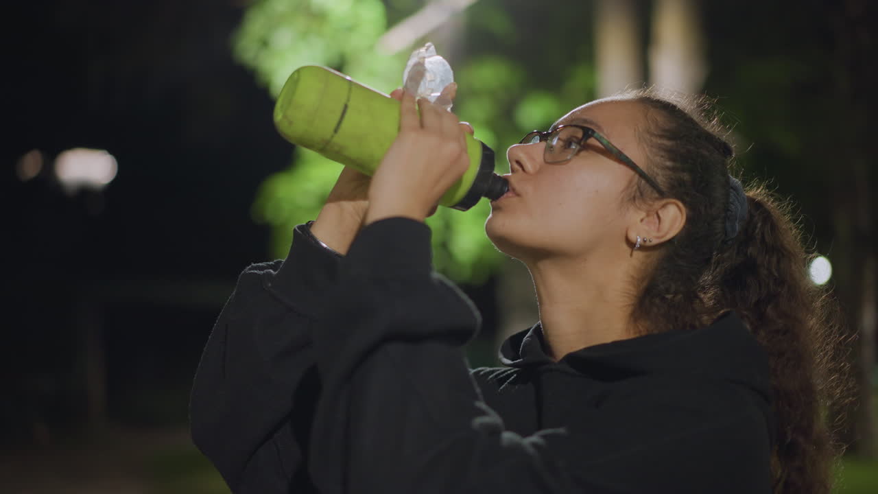 Female Pauses To Drink Beneath Luminous Trees, Woman Takes Break To Drink On Peaceful Night Path, Female Individual Refreshes With Beverage Under Streetlamp Amidst Surrounding Natural Beauty