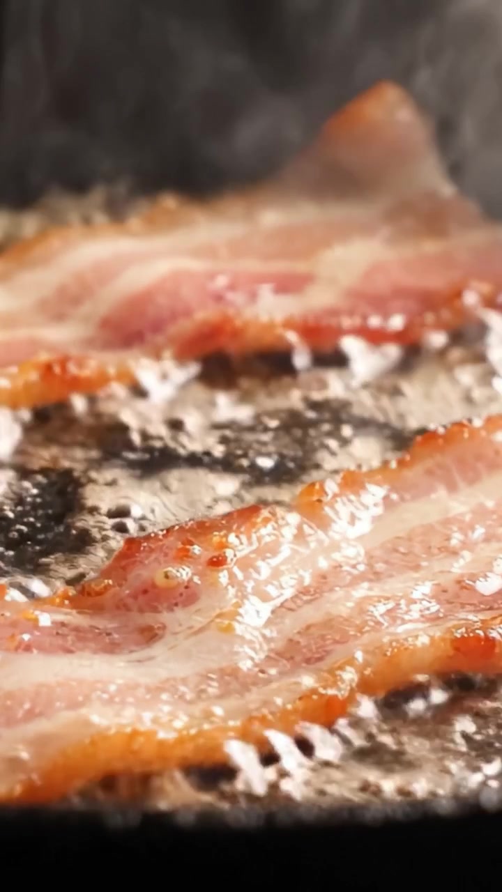 Sydende sprøde baconskiver steger på en varm pande, glinsende af olie og fedt, hvilket skaber en uimodståelig aroma og lækker knas for morgenmadsentusiaster og madelskere