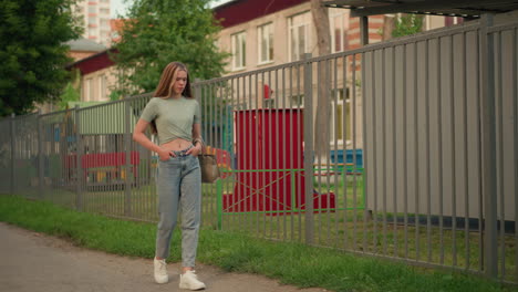 lady with hand in pocket walking along path beside iron fence, holding beige handbag, long blonde hair flowing down her back, playground structures visible through fence, green grass along walkway