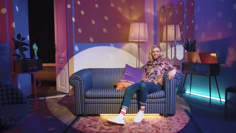 young stylish caucasian man with long hair in glasses sitting on a sofa in a retro party at home