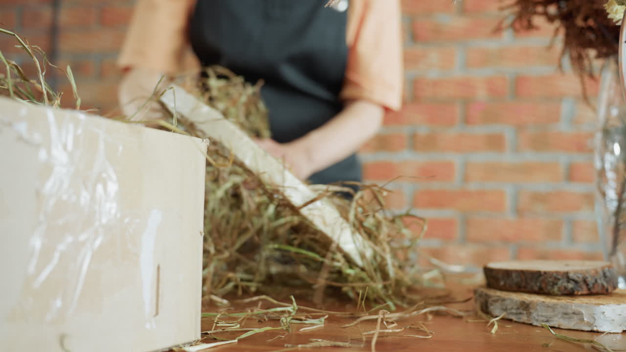 Decorator arranging hay onto wreath base on wooden table with rustic background, preparing handmade natural decoration using twine and wooden slices, focusing on eco-friendly seasonal creative design