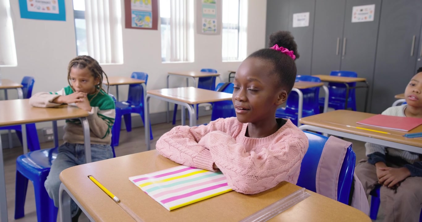 Schoolchildren watching front girl smiling replying to teacher prompt at desk with striped notebook