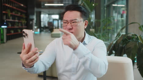 angry young man boss in shirt holding smartphone arguing over video call in modern office lobby