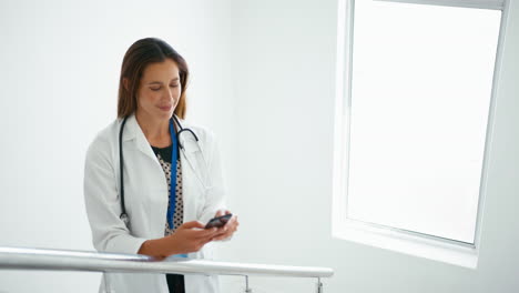 Female-Doctor-In-White-Coat-Browsing-On-Mobile-Phone-On-Stairs-In-Hospital