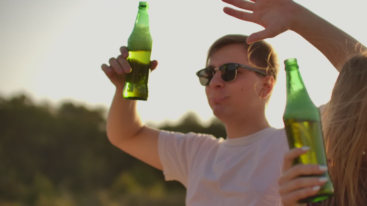 un joven brutal está bailando con gafas de sol negras de moda y una camiseta blanca con cerveza en una fiesta al aire libre con una hermosa joven rubia de cabello largo en una camisa a cuadros roja.