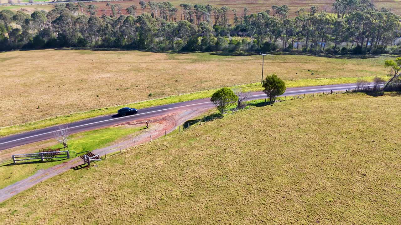 A single car travels on a winding country road through open grassy fields, captured in daylight with smooth aerial camera movement and natural lighting