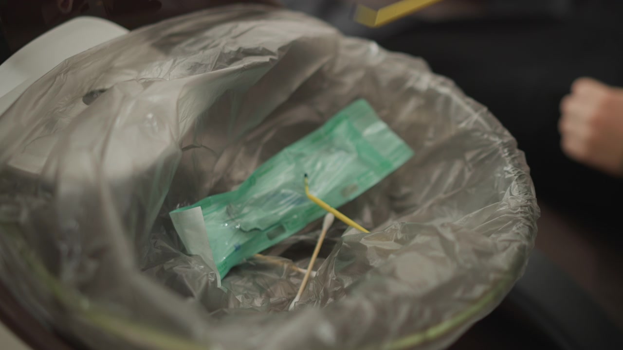 Primo piano di un cestino di plastica contenente rifiuti medici, inclusi imballaggi verdi e tamponi di cotone. La scena mostra il processo di smaltimento con movimenti minimi della telecamera, concentrandosi sugli articoli sterili