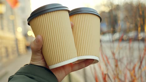 person holding with one hand two cardboard cups with coffee in the street with the light of dawn