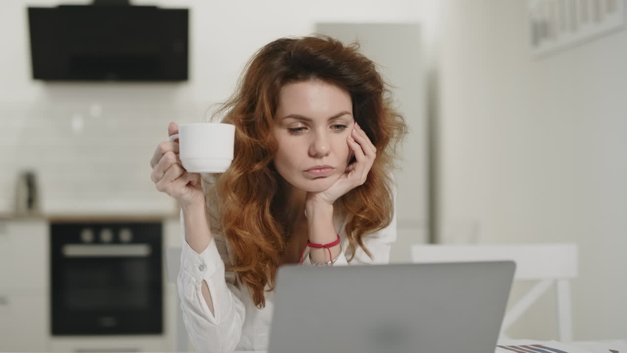 Concentrated woman drinking tea at morning kitchen. Young lady working laptop