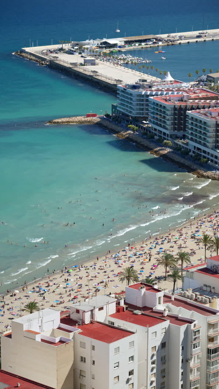 Aerial view of turquoise water, a sandy beach and seafront apartment blocks with swimmers and umbrellas visible below. Vertical