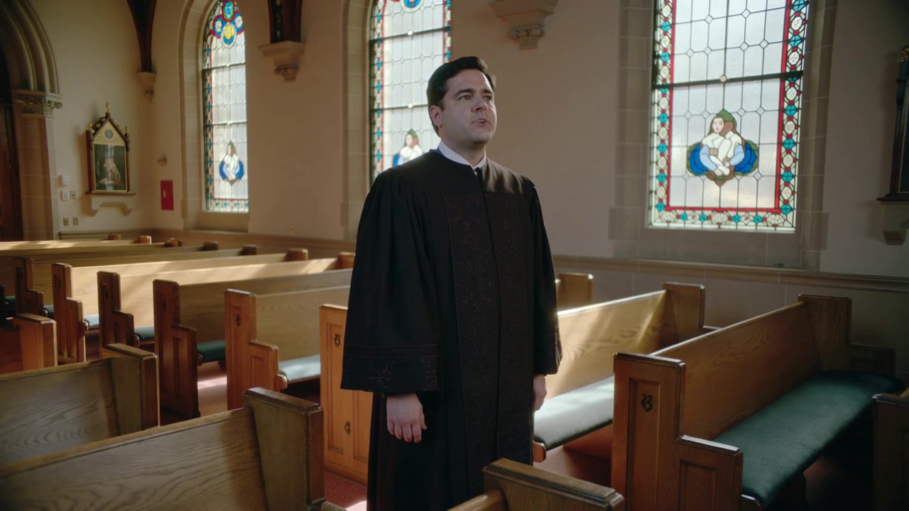 Ein Mann im Chorrock hebt im Licht der Buntglasfenster das Kinn und singt in der Kirche, mit Holzbänken.