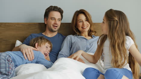 padres amistosos caucásicos felices con su hijo y su hija sentados en la cama por la mañana