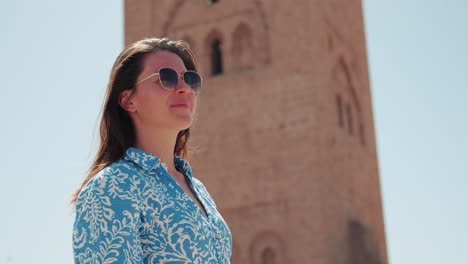 Detail-of-young-caucasian-woman-in-dress-with-sunglasses-smiling-and-looking-around-in-front-of-Koutoubia-Mosque,-Marrakech,-Morocco