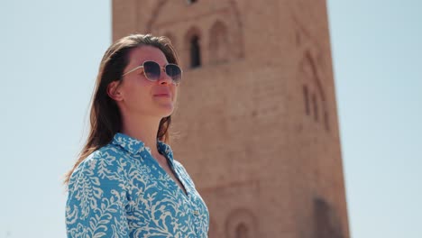 detalle de una joven caucásica vestida con gafas de sol sonriendo y mirando a su alrededor frente a la mezquita de koutoubia, marrakech, marruecos
