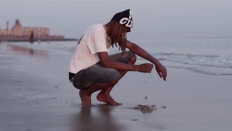 Ein-Afrikanischer-Mann,-Männlicher-Mann,-Modelljunge,-Sitzt-Auf-Kniebeugen-Am-Strand,-Meeresufer,-Meerwasser-Im-Sonnenuntergang-Und-Berührt-Sand