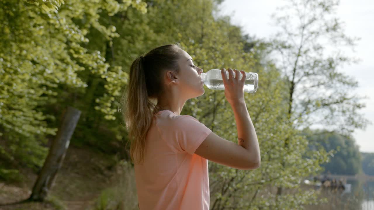 mujer bebiendo agua de una botella de pie junto al lago y al bosque