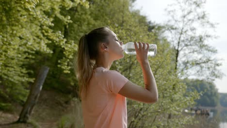 Woman-drinking-water-from-bottle-standing-by-lake-and-woods