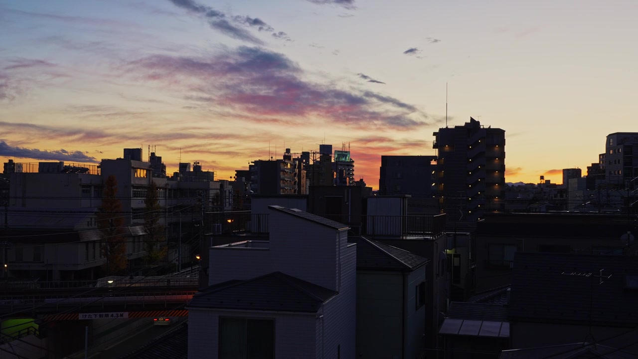 Timelapse atmosférico de una ciudad japonesa urbana mientras nubes coloridas de atardecer cruzan el cielo y la luz del día transiciona al crepúsculo, con trenes de cercanías pasando por las vías férreas debajo