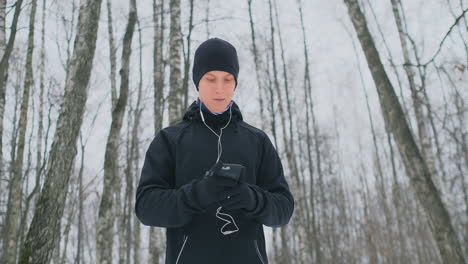 Ein-Junger-Mann-In-Einem-Schwarzen-Auslöser-Bereitet-Sich-Im-Winter-Auf-Das-Joggen-Im-Park-Vor,-Läuft-Mit-Musik-Auf-Dem-Smartphone-Und-Läuft-Mit-Einem-Sprachassistenten-Durch-Den-Wald,-Um-Geschäfte-Zu-Machen-Und-Einen-Gesunden-Lebensstil-Zu-Führen