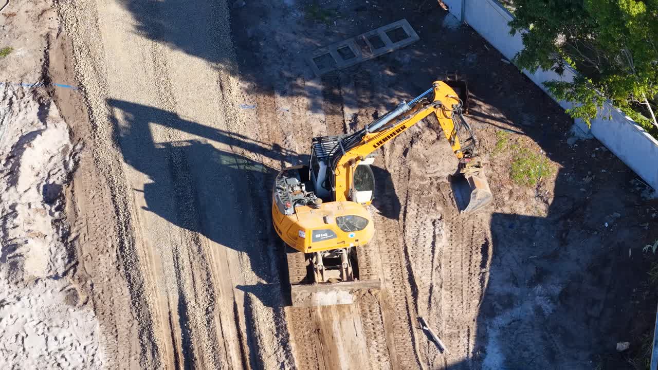 Yellow excavator moves and loads dirt at sunny construction site, captured by overhead drone shot