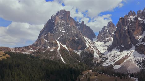 una vista impresionante de la montaña sassolungo, con picos cubiertos de nieve y un denso bosque de pinos en las dolomitas, bajo un cielo azul brillante con nubes esponjosas