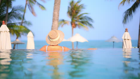 Espalda-De-Una-Mujer-Hermosa-Con-Sombrero-Para-El-Sol-Disfrutando-De-La-Vista-Desde-El-Interior-De-La-Piscina-Infinita-Apoyada-En-El-Borde-En-Un-Resort-De-Isla-Tropical,-En-Cámara-Lenta