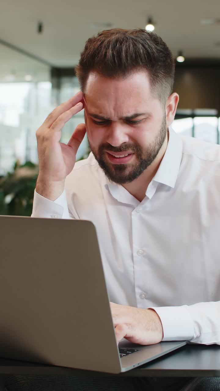 Tired ill young businessman using laptop suffering from headache problem tension in hotel lobby