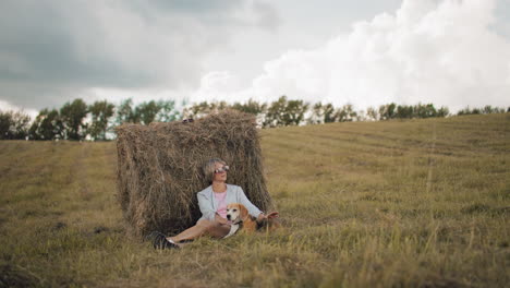 woman wearing sunglasses and casual outfit sits against hay bale in open farmland, gently petting her dog, golden grass, rolling hills, and cloudy sky