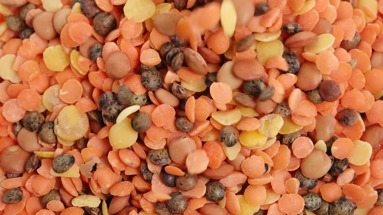 A variety of lentils and beans cascade onto a surface, creating a vibrant, textured pattern in bright lighting