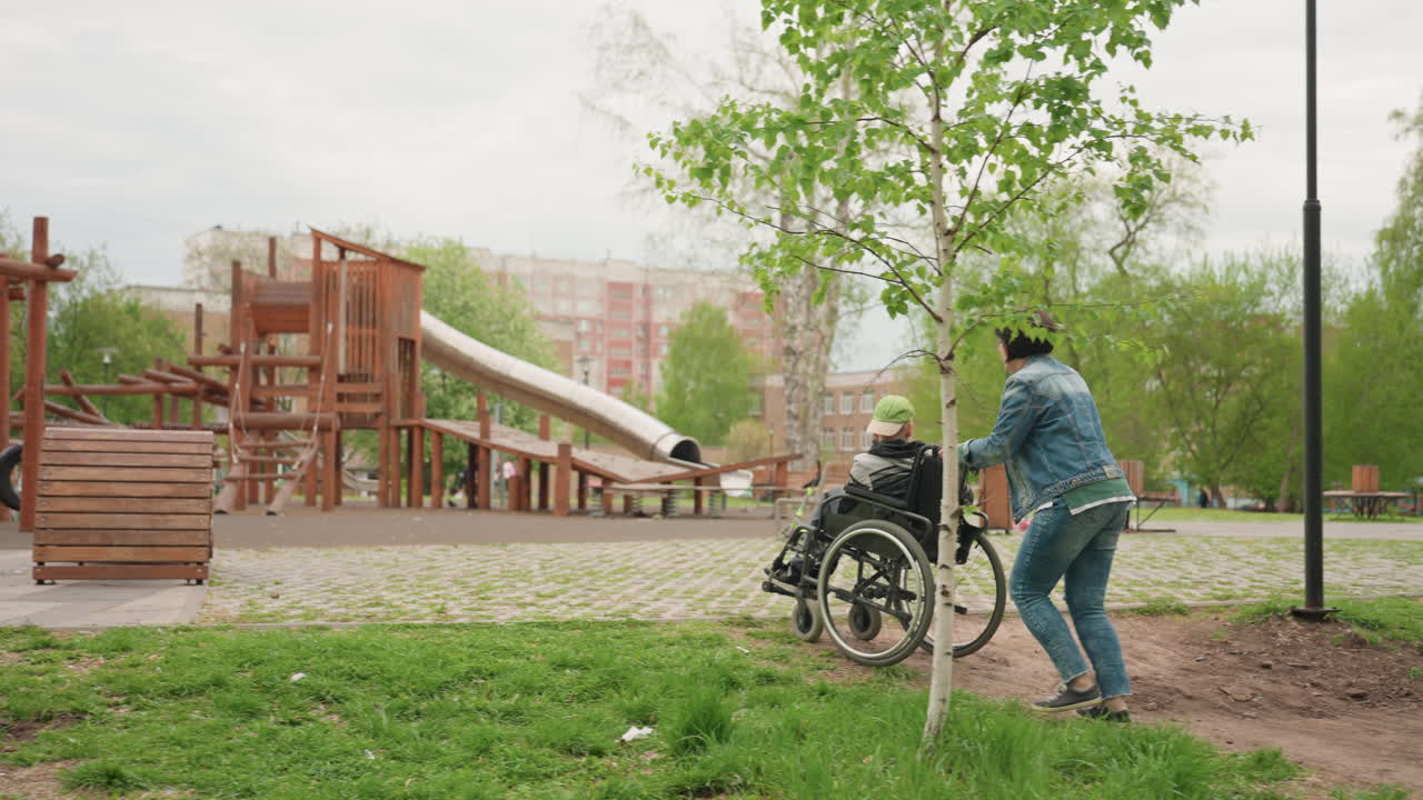 Friend Guides Disabled Child Outdoors, Mobility Aid User Explores Park With Friend, Person Using Wheelchair Engages With Playground Equipment Alongside Attentive Caregiver And Friends