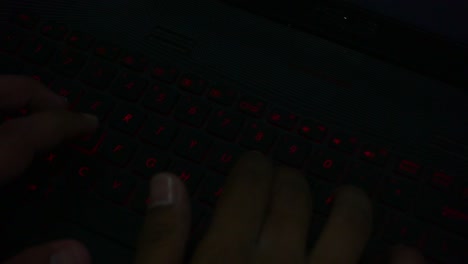 Vista-Superior-De-La-Mano-Escribiendo-En-El-Teclado-De-Iluminación-Roja-De-La-Computadora-Portátil-En-La-Habitación-Oscura,-Primer-Plano