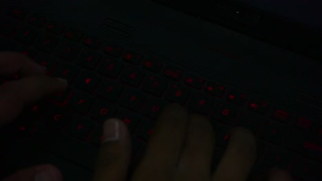 vista superior de la mano escribiendo en el teclado de iluminación roja de la computadora portátil en la habitación oscura, primer plano
