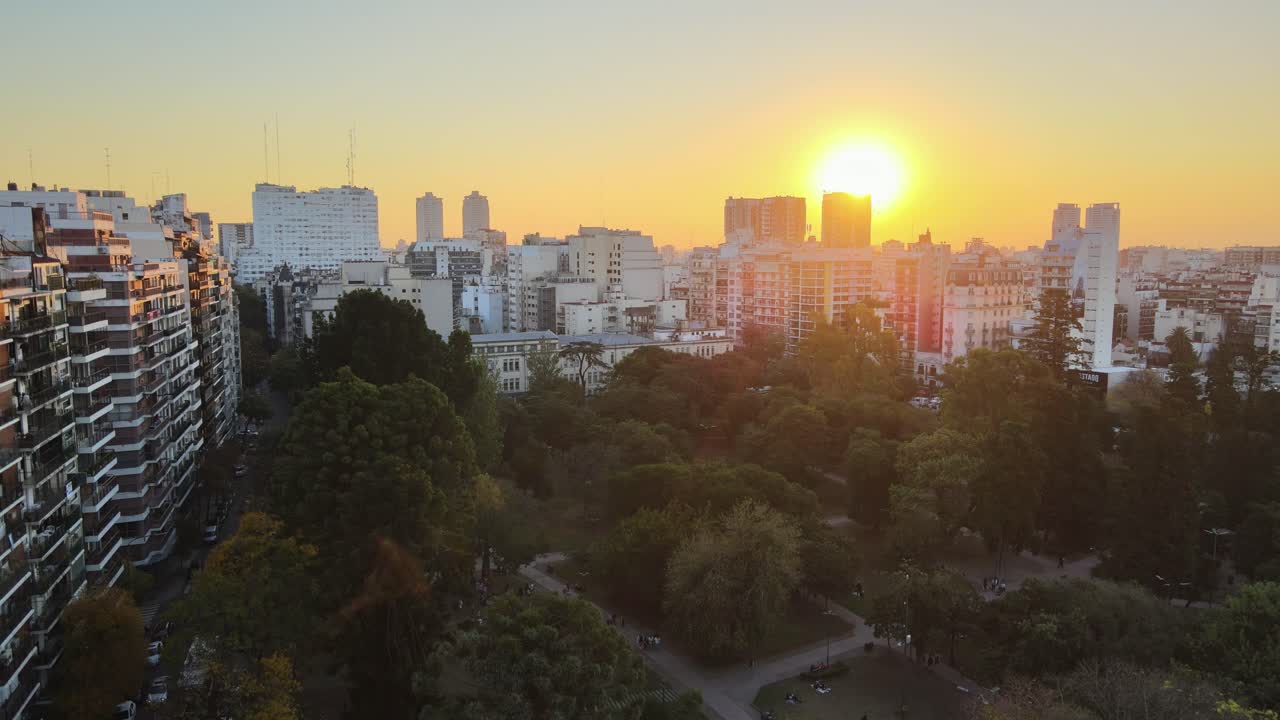 Descending aerial view over Parque Rivadavia’s treetops with sunlit buildings and sunset in the Buenos Aires skyline, Argentina