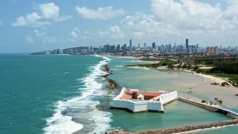 Dolly-out-aerial-drone-shot-of-the-historic-star-shaped-Reis-Magos-fort-built-on-a-reef-with-the-coastal-capital-city-of-Natal-in-Rio-Grande-do-Norte,-Brazil-in-the-background-on-a-warm-summer-day