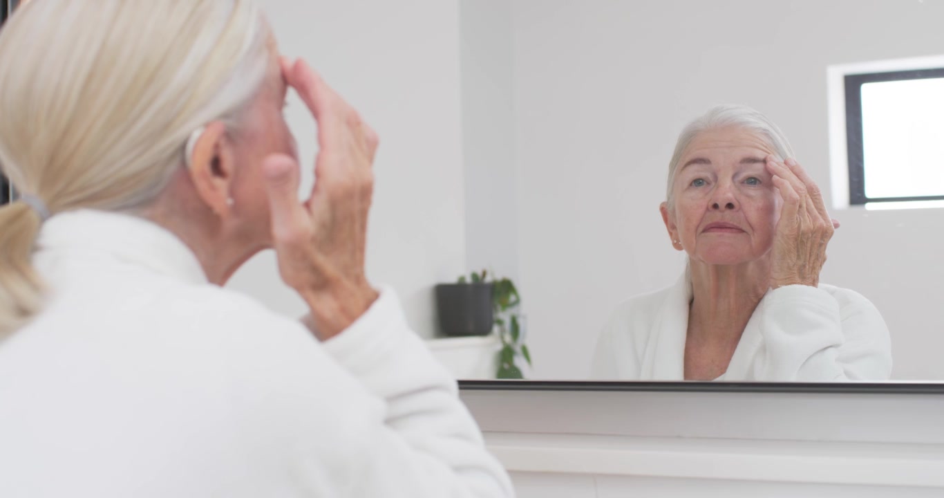 Una donna anziana, notando le rughe nello specchio, applica la crema contorno occhi alla toeletta, indossando una vestaglia bianca e con un apparecchio acustico.