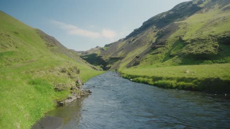 A-small-river-and-mossy-hills-in-Iceland,-sunny-green-rocks-and-blue-water-in-the-foreground