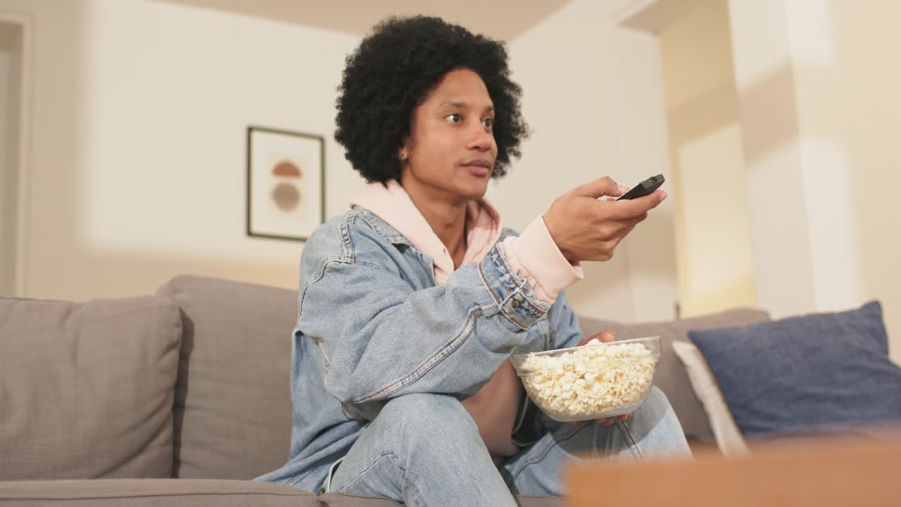 Uomo afroamericano che cambia canale con il telecomando in salotto, mangiando popcorn, alla ricerca di un nuovo programma