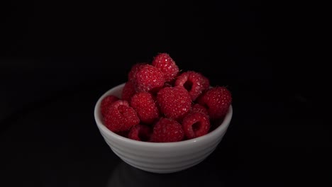 pile of red ripe raspberries in small white bowl revolves on black background.