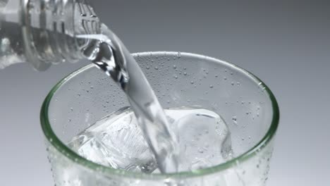 slow motion pouring clear fizzy drink from glass bottle into chilled glass with ice