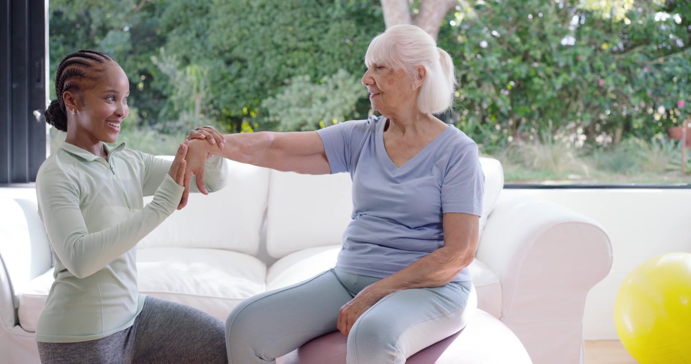 Sportswear trainer guiding Senior extending right arm for rehab, doing wrist turns on ball at home