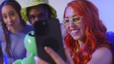 fotografía de estudio de un grupo de amigos de la generación z sentados en el sofá posando para una selfie con un alienígena de juguete en el teléfono móvil 6