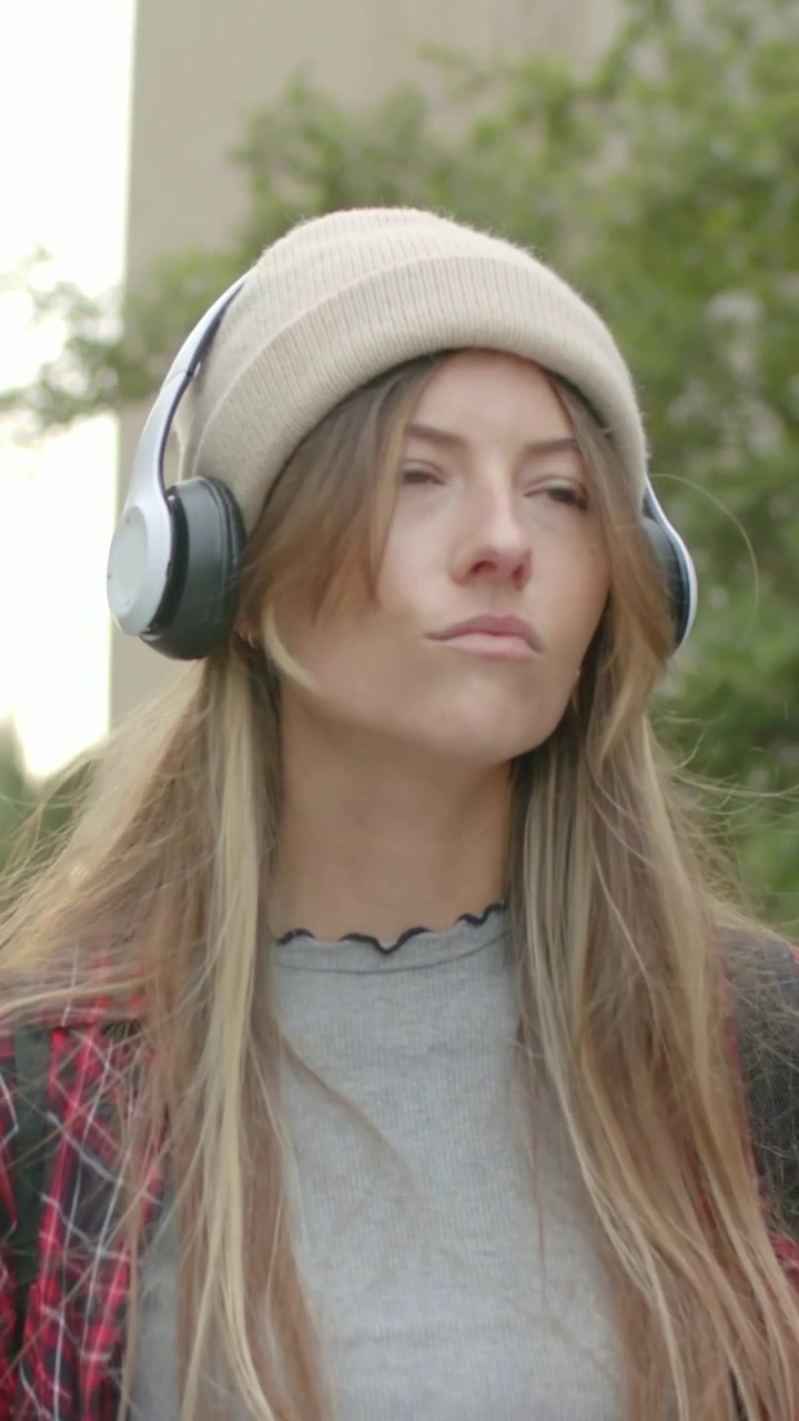 Video verticale: Musica che parte, donna che mima le parole su un sentiero del campus, indossando cuffie e berretto.