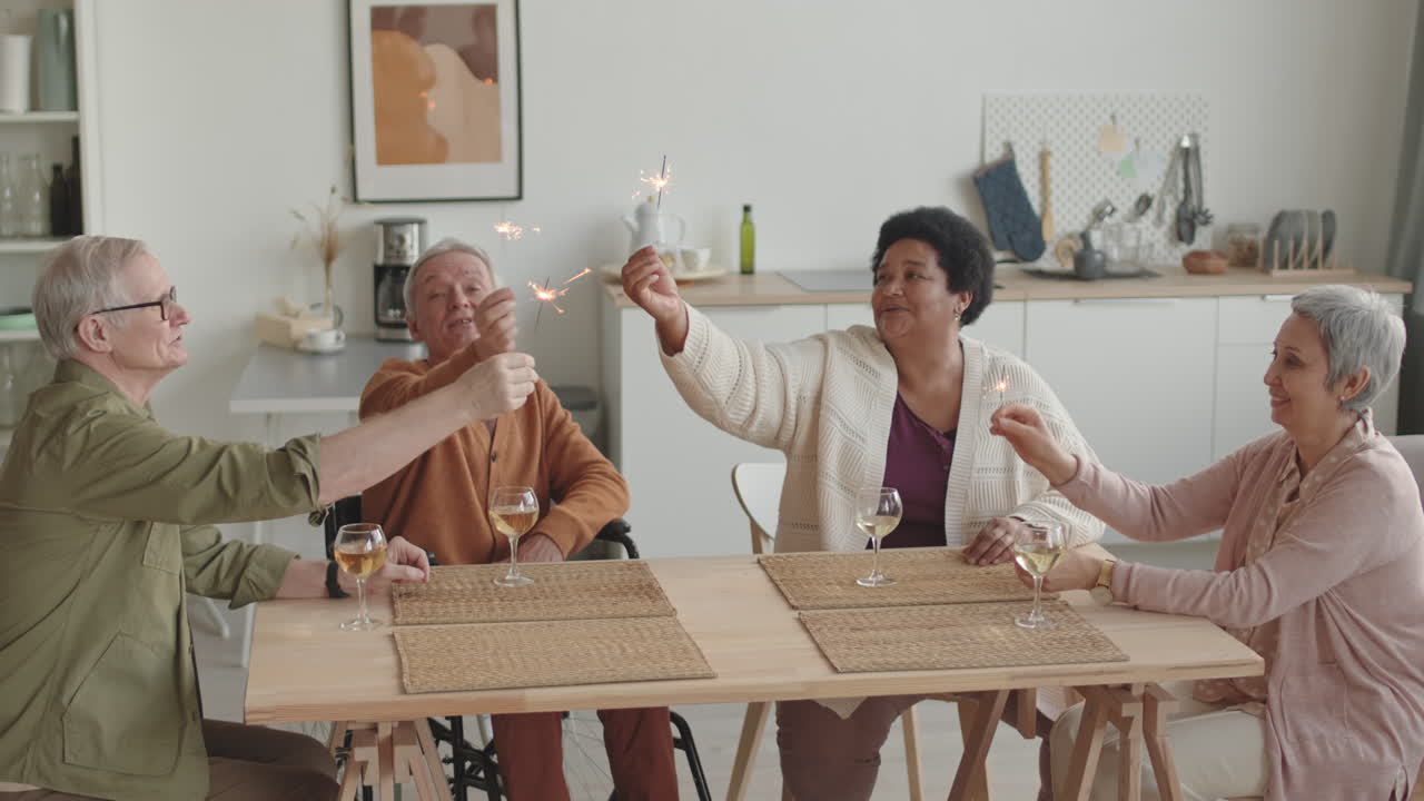 Medium long of Asian and African women, senior handicapped and Caucasian men sitting at table in dining room, holding lit Bengal Lights smiling