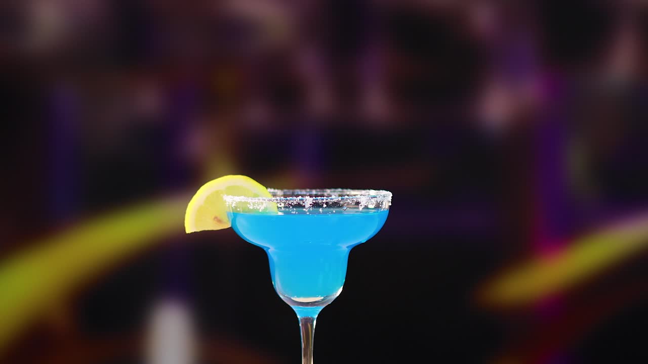 A hand presents a vibrant blue margarita cocktail garnished with a lemon slice against a blurred, colorful background