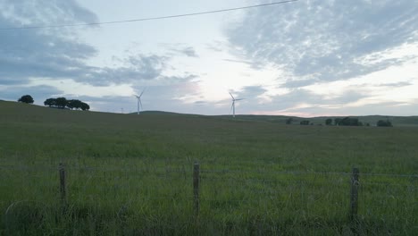 Fenceline-dolly-shot-past-spinning-wind-turbines-in-morning-pasture