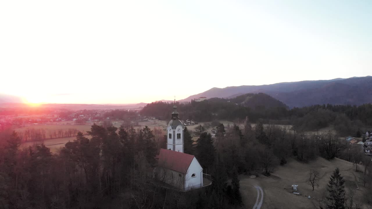 Aerial flight at the same height of the trees that surround the church. Drone flying over a Slovenian church at Sunrise.