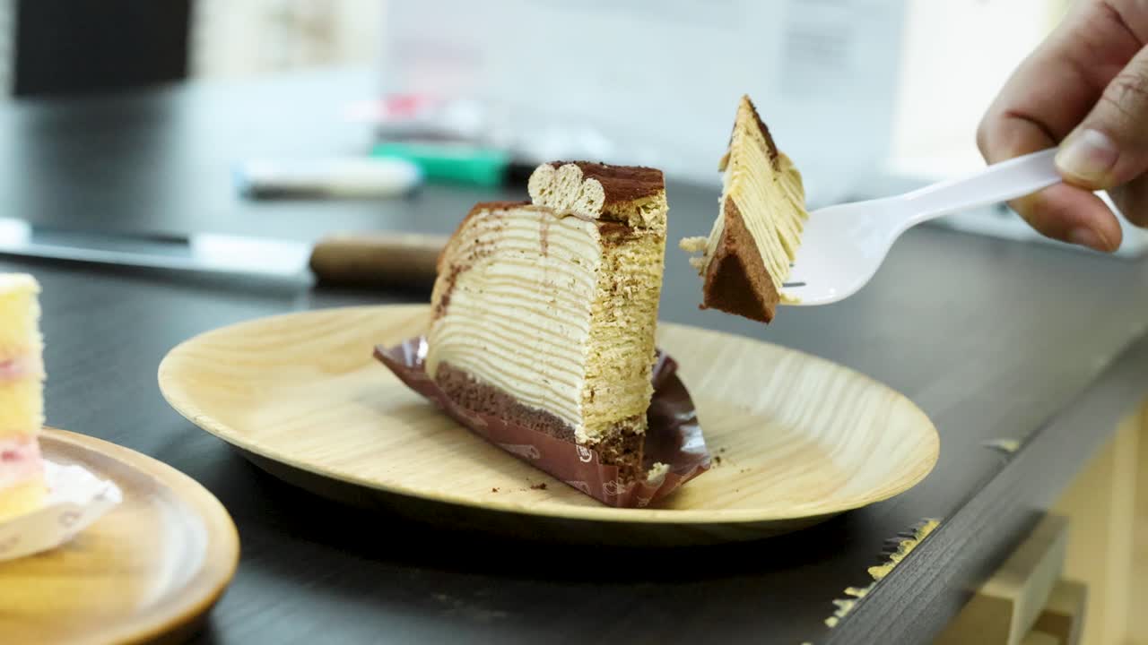 Hand uses plastic fork to cut and lift tiramisu crepe cake slice indoors
