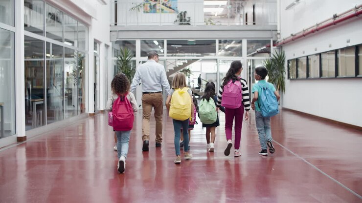 Vista trasera de niños caminando con un tutor masculino en el pasillo de la escuela.