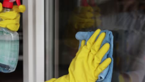 woman, housework and housekeeping concept - woman in gloves cleaning window with rag and cleanser spray at home