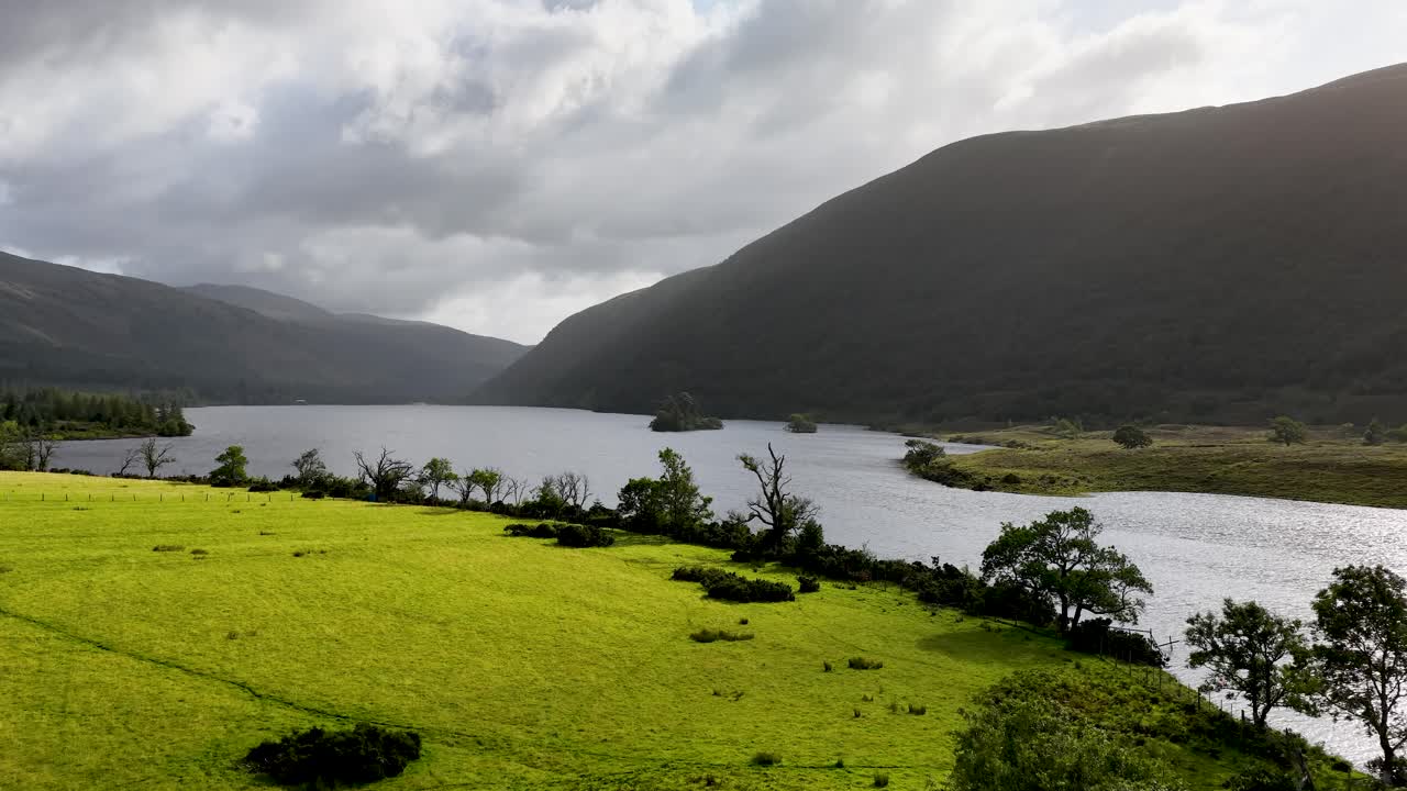 Drone pans across lush green valley, reflective lake, and mountains under dramatic cloudy skies